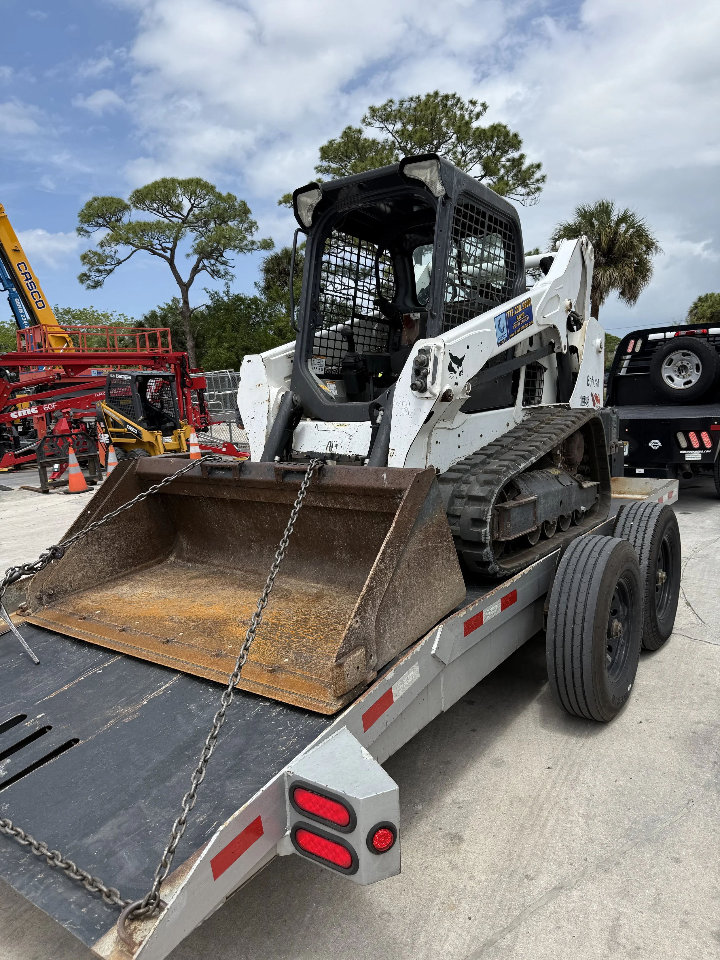 A white skid-steer loader with black protective cage on a flatbed trailer with chains securing it, and orange construction cones in the background.