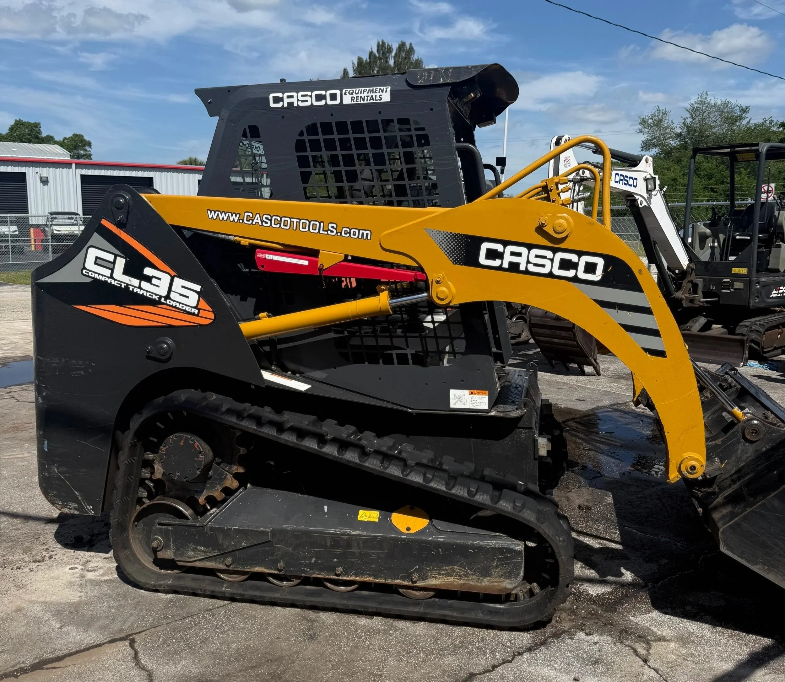 Yellow and black construction skid steer loader with the brand name CASCO and model CL36, parked outdoors under a blue sky with some clouds.