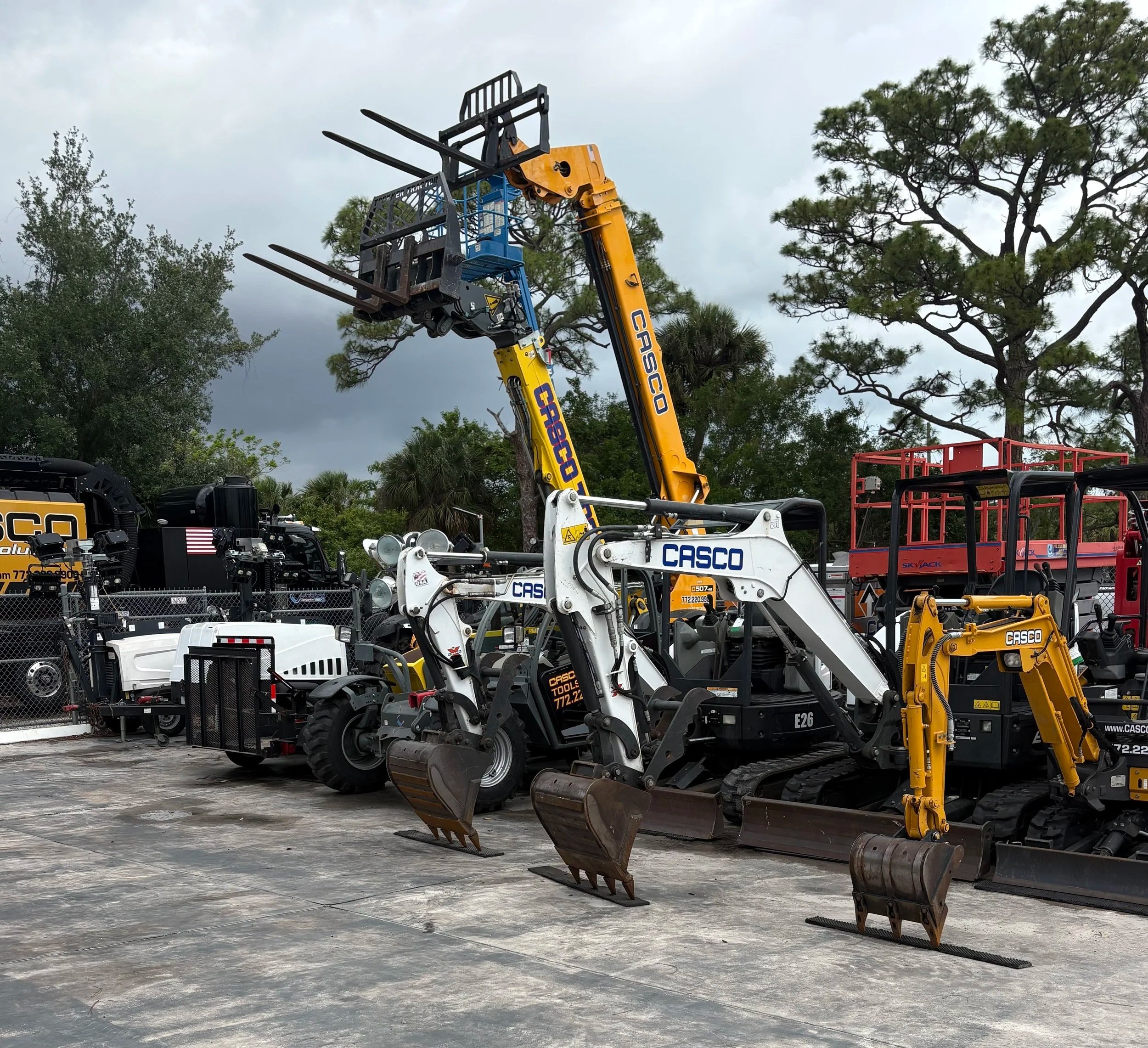 Construction equipment including multiple forklifts and mini excavators parked outdoors, with trees and a cloudy sky in the background.
