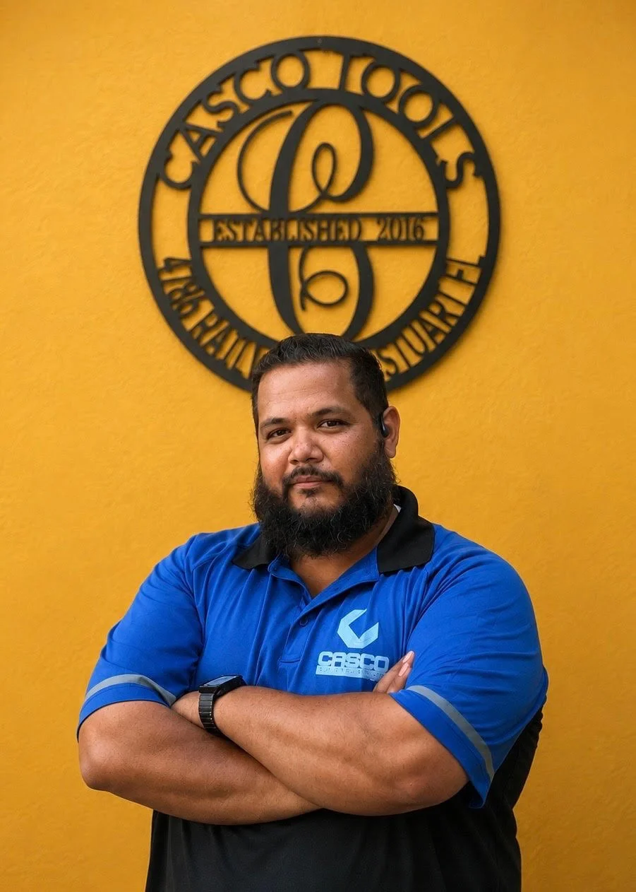 A man with a beard wearing a blue CASCO shirt and a watch, standing with arms crossed in front of a yellow wall with a black circular ASCO logo.