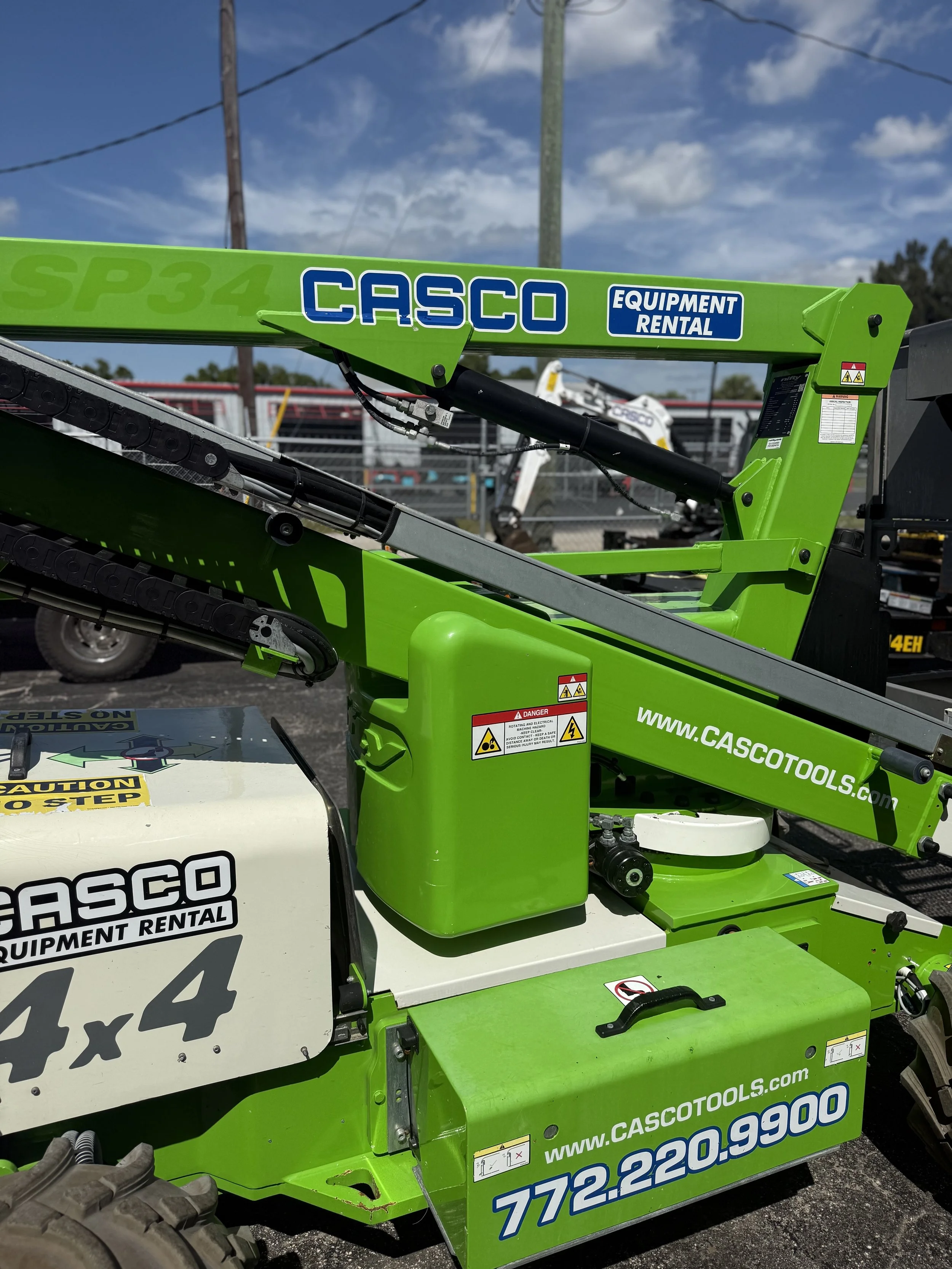 Green Casco equipment rental lift with hydraulic arm and chains, parked outdoors with a blue sky and power lines in the background.