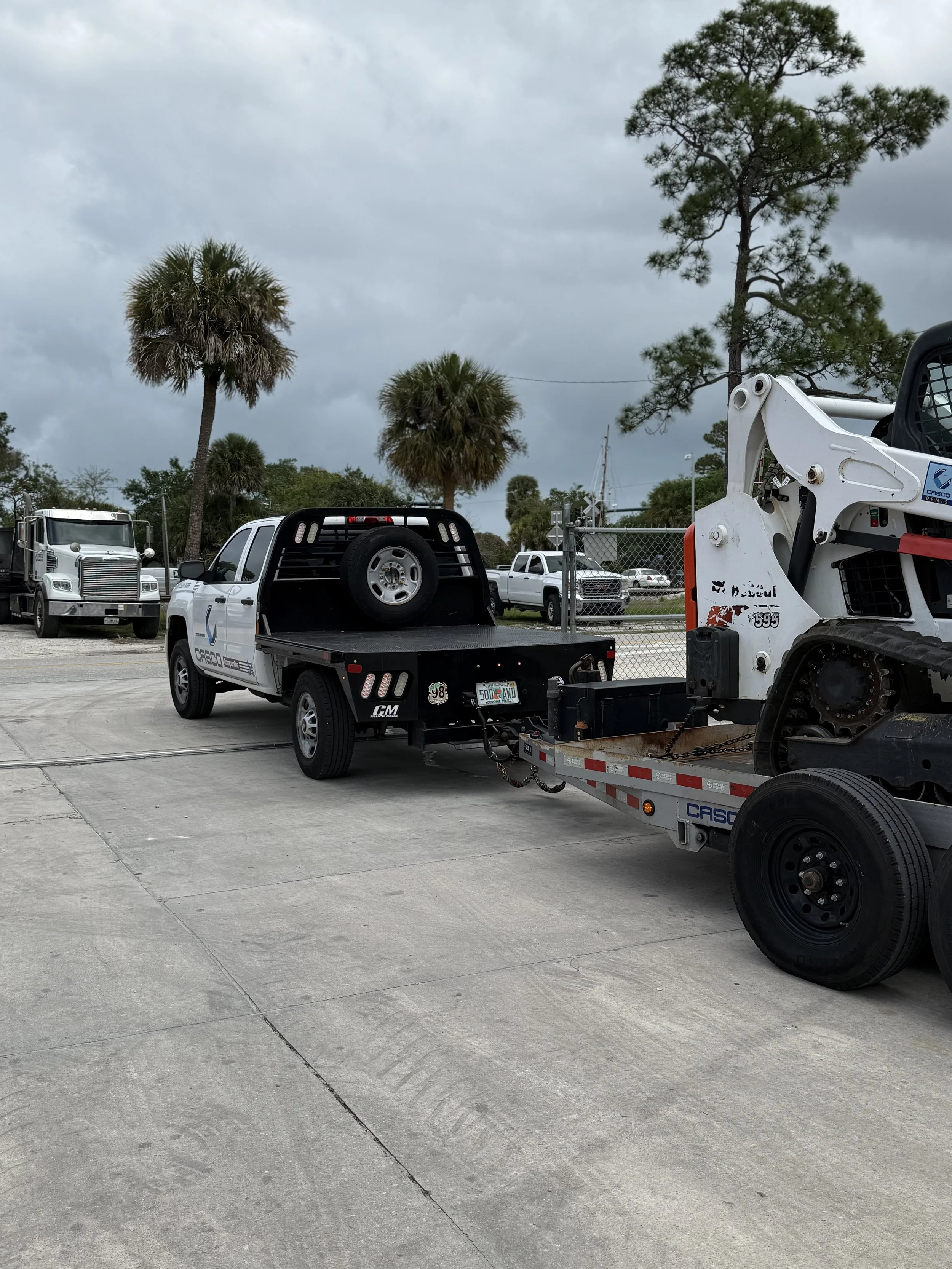 A white pickup truck with a flatbed carrying a small excavator, parked on a concrete lot with palm trees and other parked vehicles in the background.