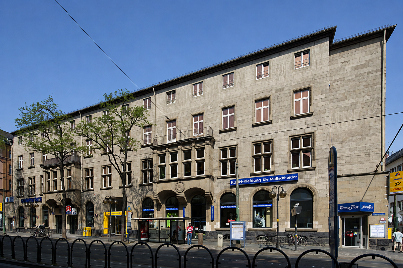 Mehrstöckiges Gebäude mit Geschäftsläden im Erdgeschoss, Bäumen davor, Straßenbahnlinien und Menschen auf der Straße, blauer Himmel.