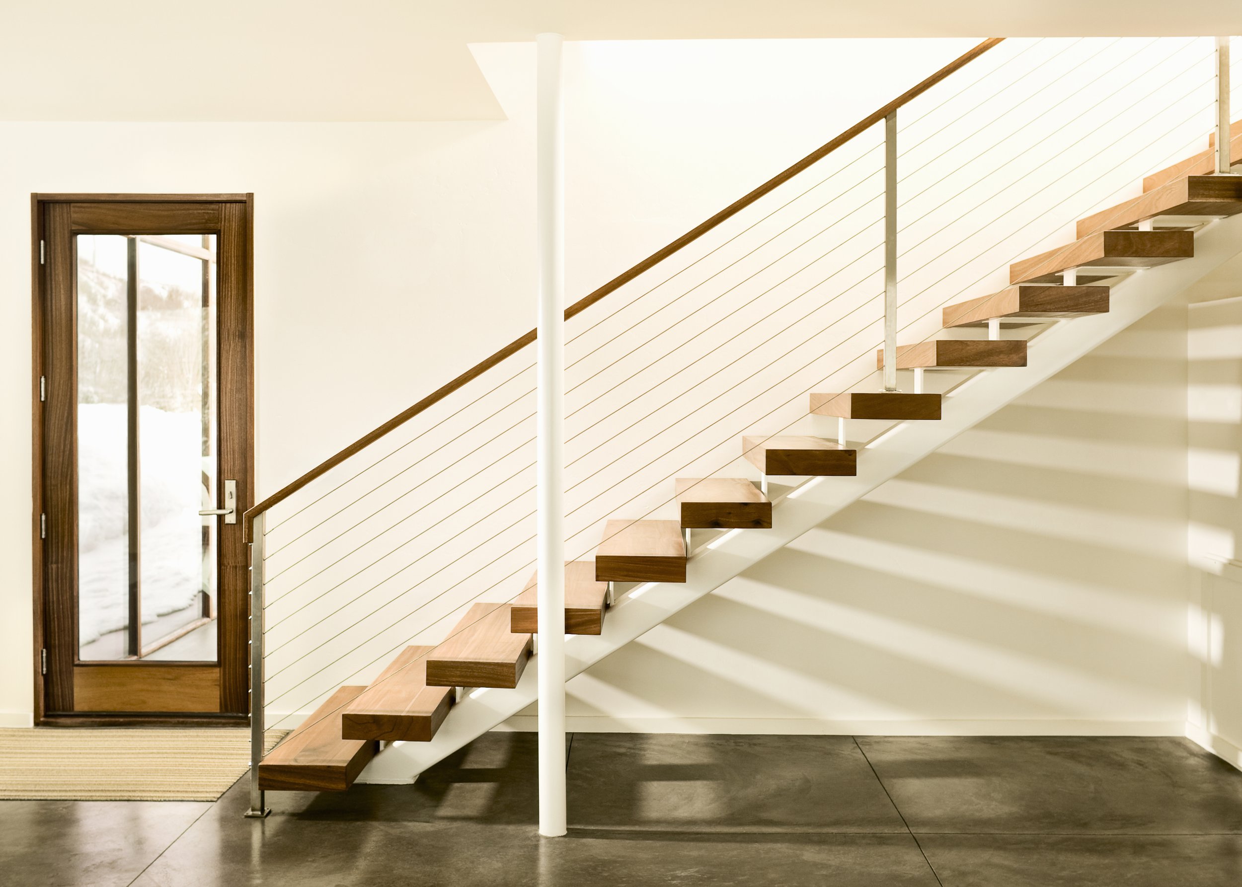 Interior view of a modern staircase with wooden steps, white support structure, and a wooden handrail, leading to an upper floor in a house with a wood-framed glass door nearby.