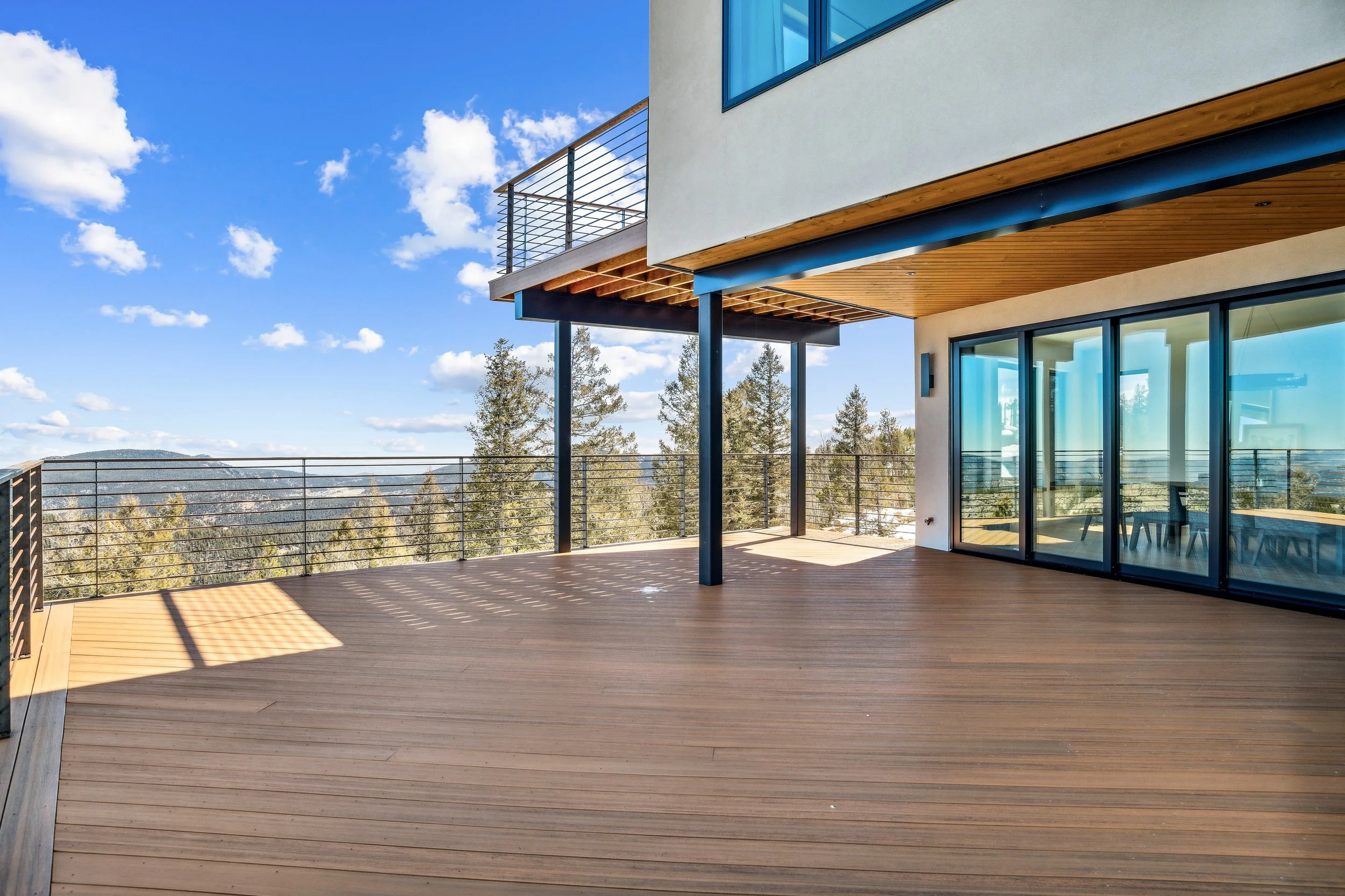Modern balcony with wooden flooring, glass sliding doors, and a view of trees and mountains under a blue sky with scattered clouds.