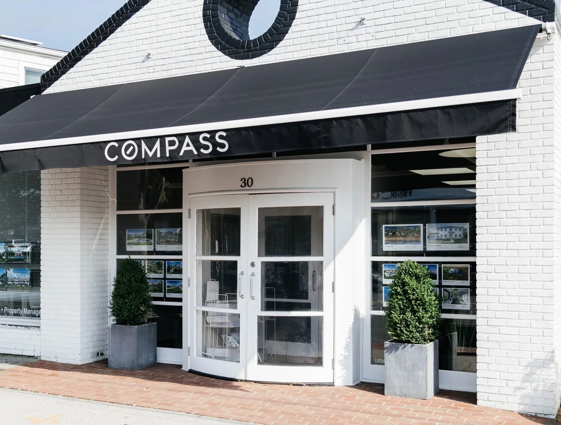 Exterior of a commercial property with a black awning and white brick facade, displaying the name 'COMPASS'. The entrance has glass double doors, flanked by two small potted plants, and windows showcasing property listings.