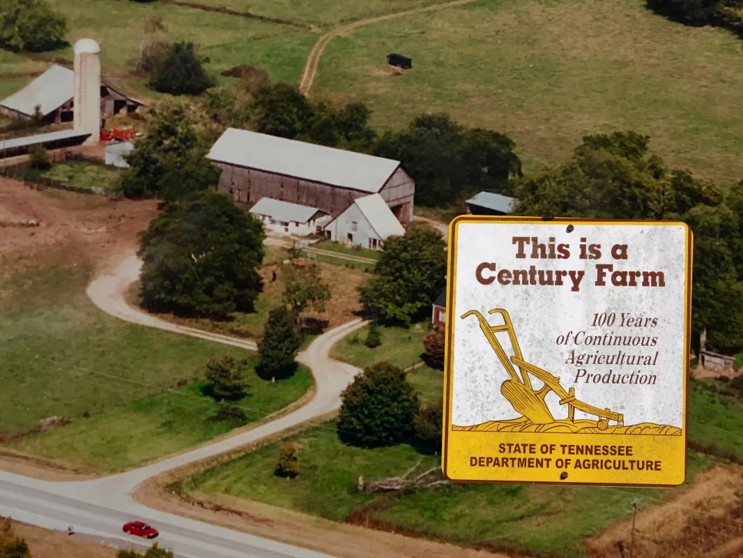 Aerial view of a historic farm with a barn, silo, and farm buildings, surrounded by greenery, with a sign in the foreground that reads 'This is a Century Farm, 100 Years of Continuous Agricultural Production, State of Tennessee Department of Agriculture.'
