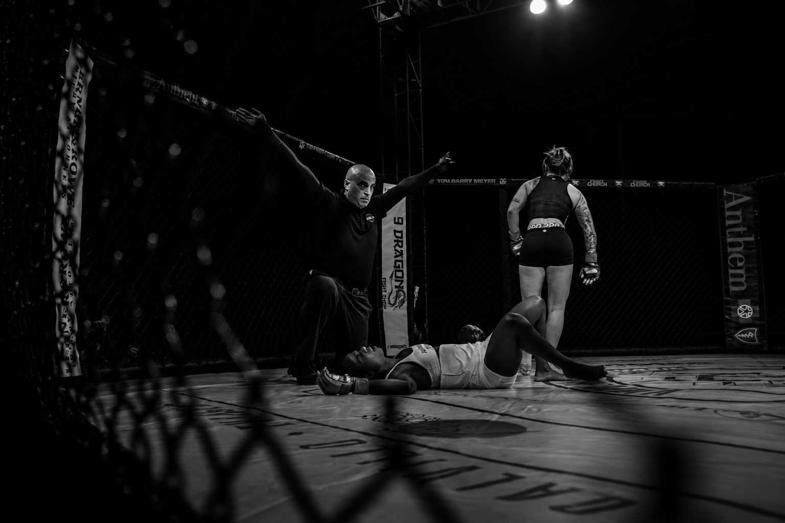 Mixed martial arts fighter lies on the cage floor, possibly injured or exhausted, while another fighter is seen walking away in the background. An official stands nearby in the octagon.