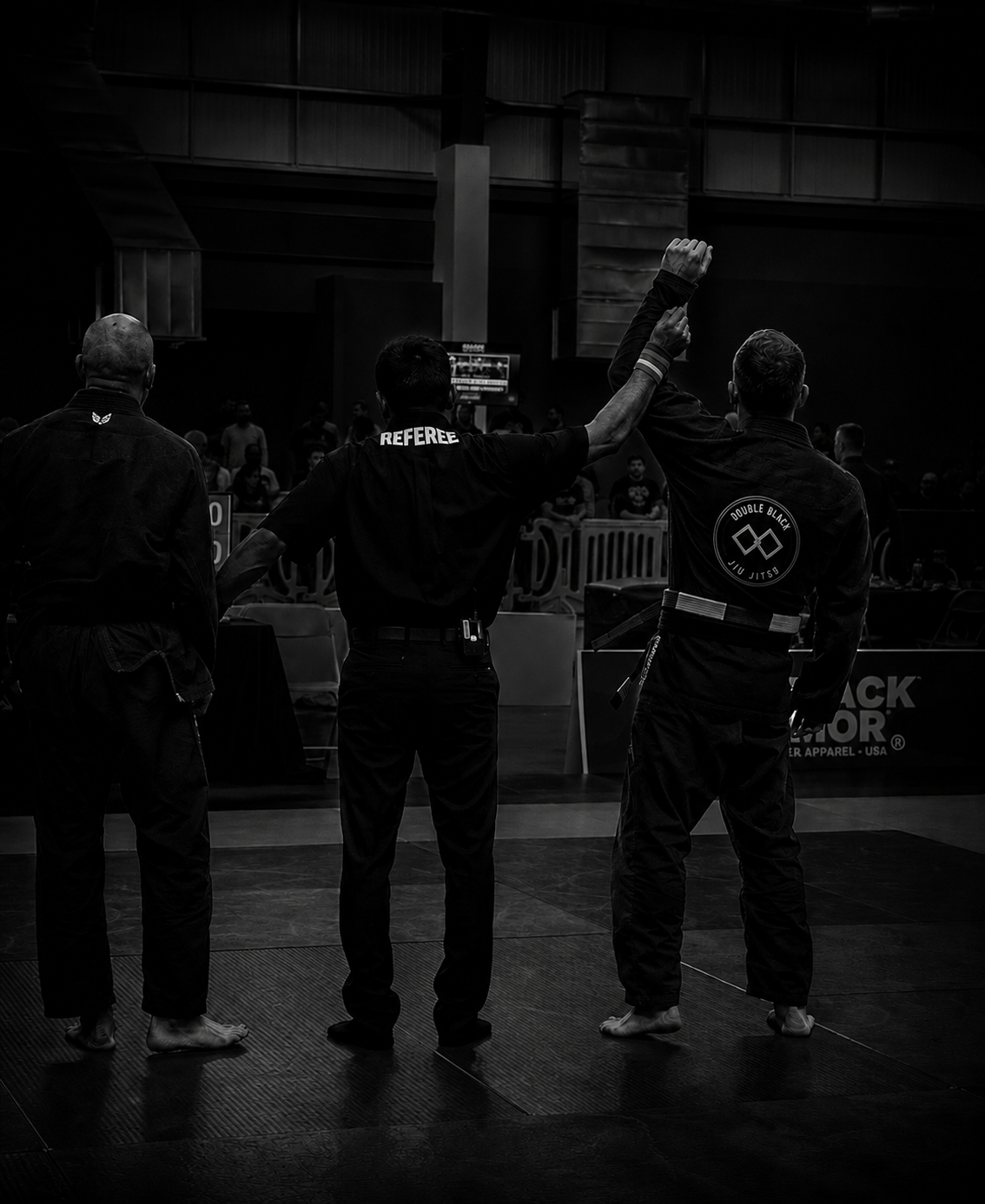 A referee signals the winner in a Brazilian Jiu-Jitsu competition while two competitors stand with their backs to the camera. One competitor has a black gi with a patch that says "Double Black" and a black belt, the other has a black gi with a butter