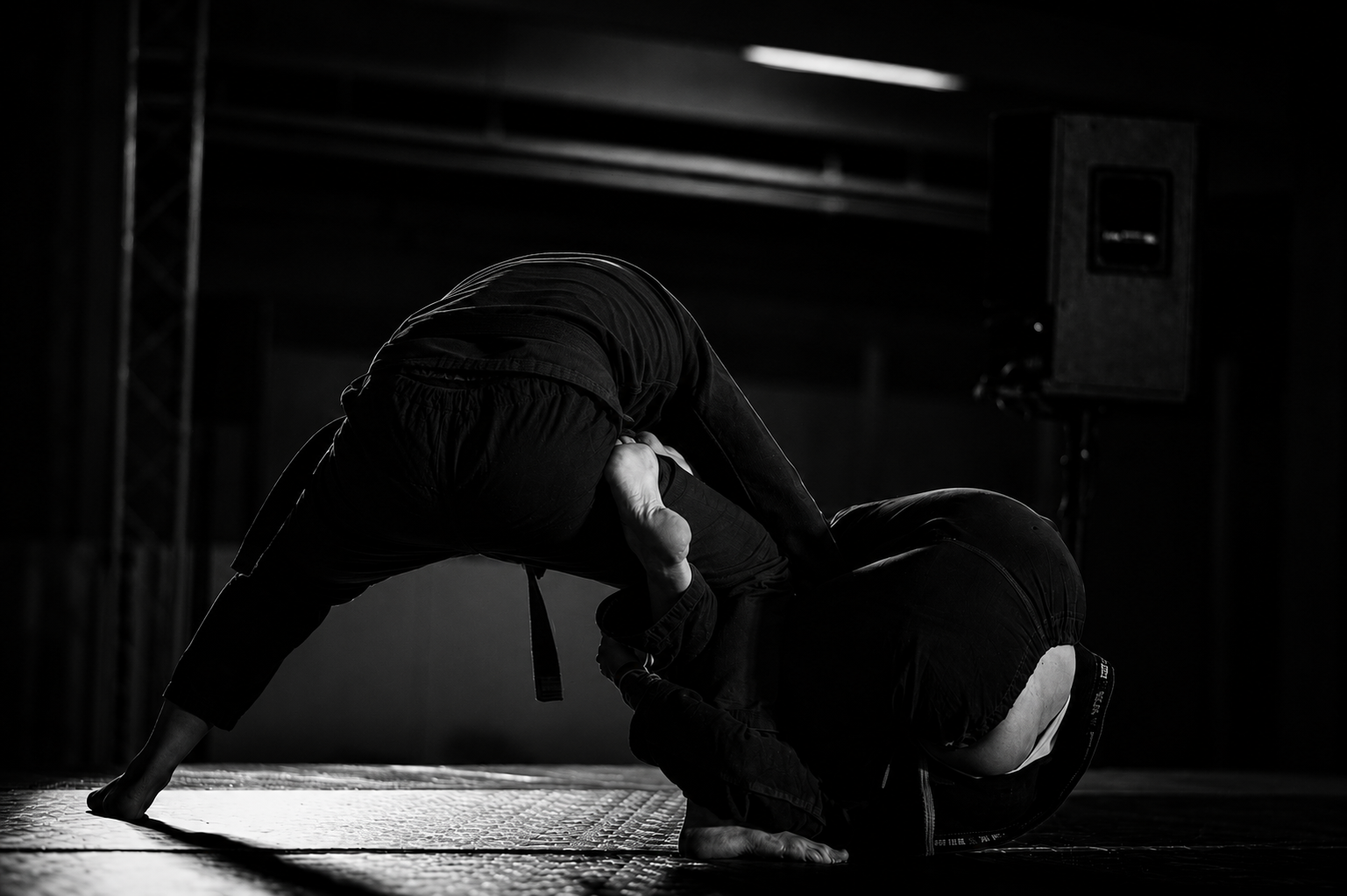 Two mixed martial arts fighters in black gi grappling on a mat in a dimly lit training area with a speaker in the background.