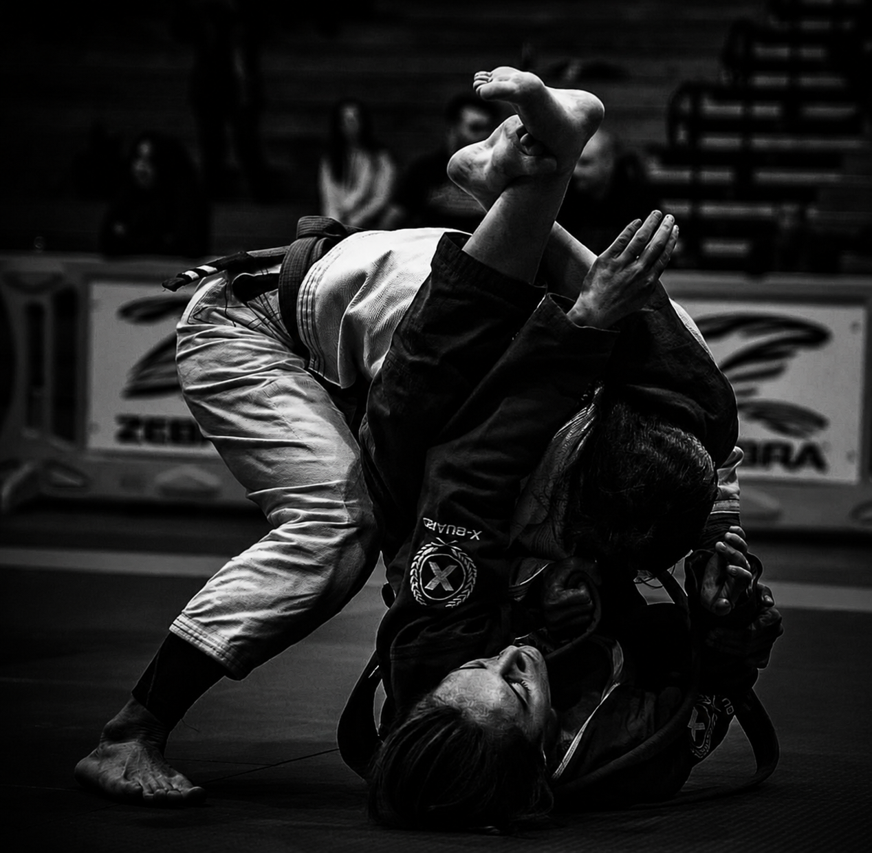 Two women practicing Brazilian Jiu-Jitsu on the mat, one in a black gi and the other in a white gi, with the woman in black executing a submission hold and the woman in white attempting to defend.