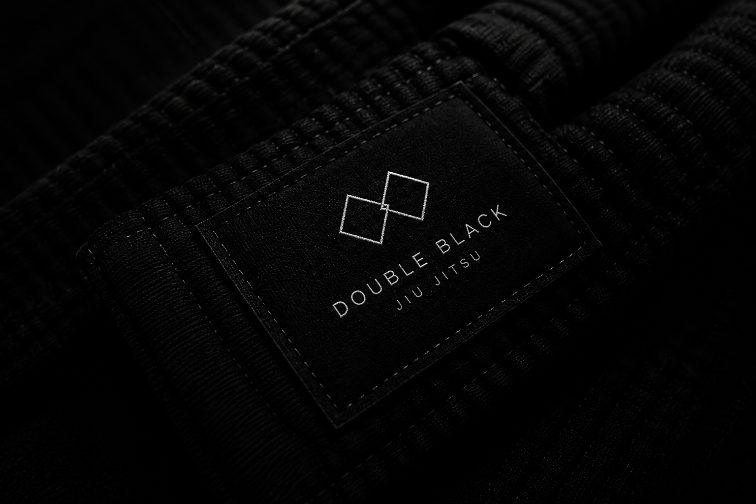 Close-up of a black martial arts gi with a leather patch reading 'Double Black Jiu Jitsu' and a minimalist logo of two overlapping squares.