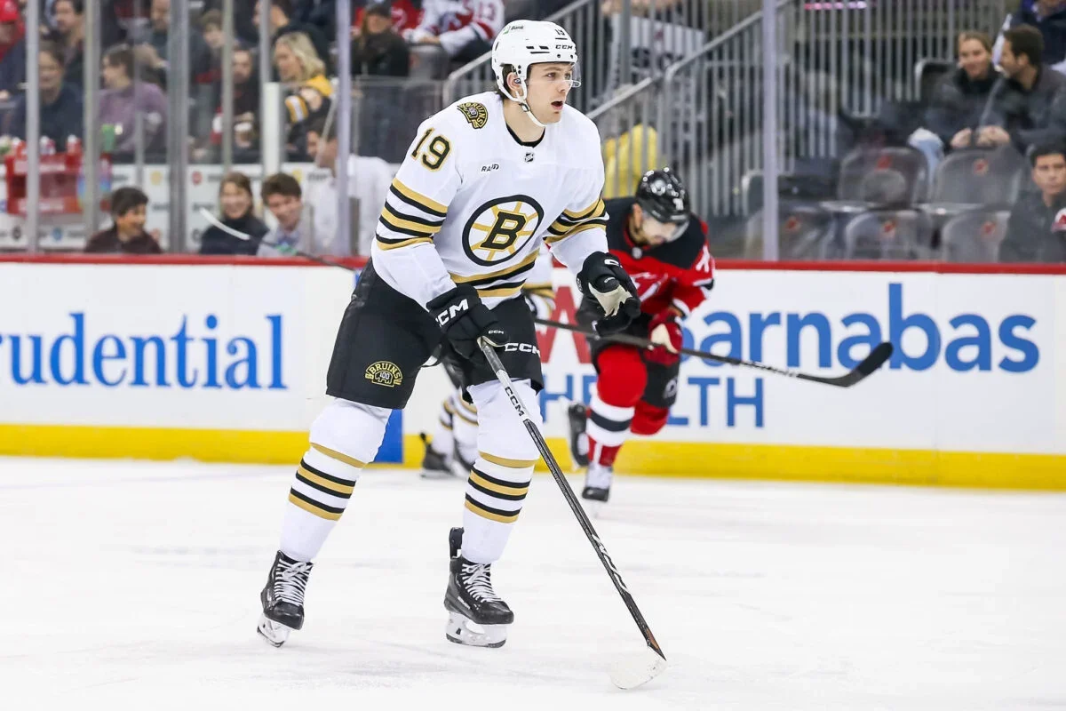An ice hockey player wearing a white jersey with black and yellow accents, number 19, and the Boston Bruins logo, is controlling the puck on the ice during a game. There are spectators in the background watching the game.