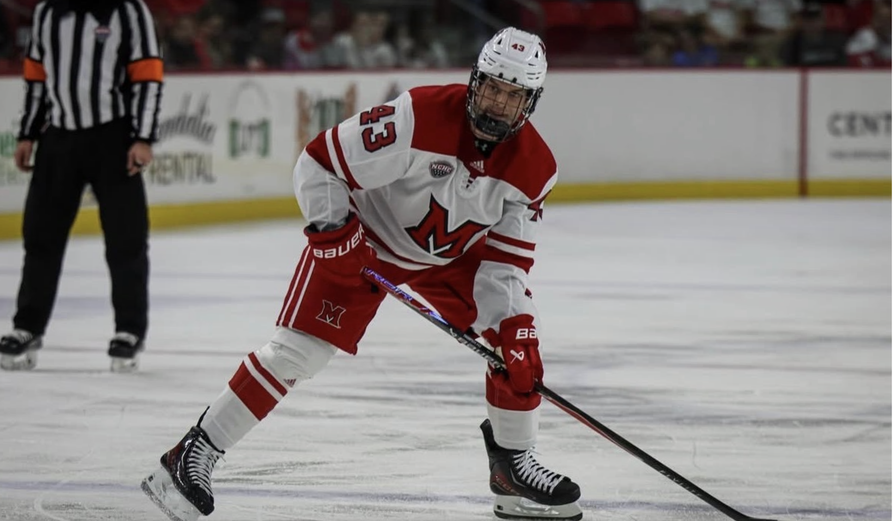 Ice hockey player in red and white uniform with the letter M on the front, bent forward on ice, holding a hockey stick, wearing a helmet and skates, with a referee in the background and spectators watching from the stands.