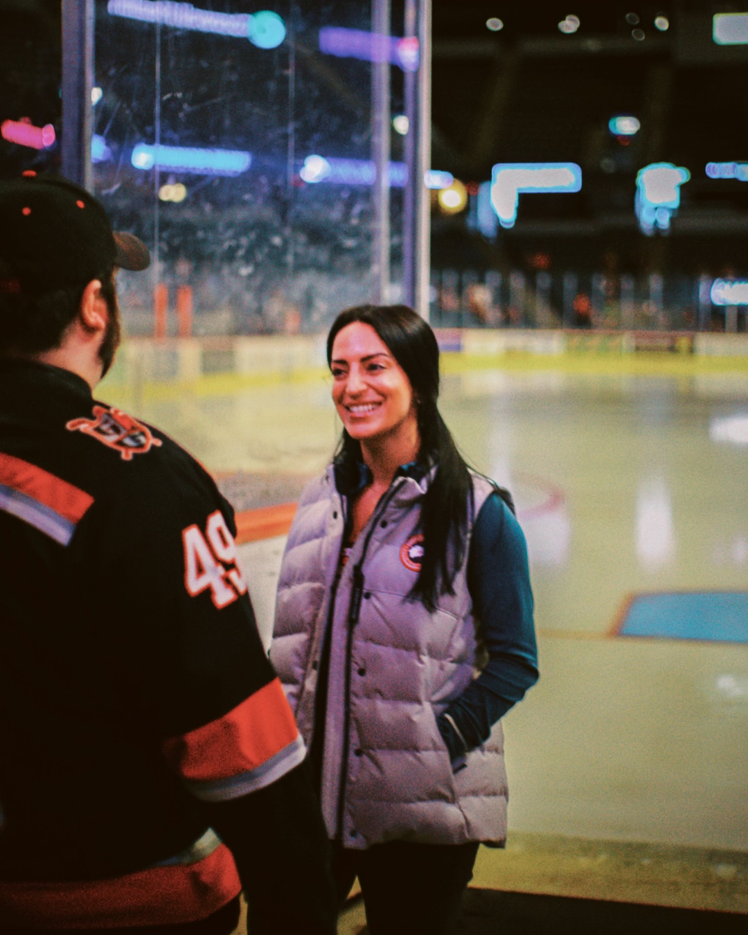 A woman smiling and talking to a man in a hockey rink, with the rink and bokeh lights in the background.