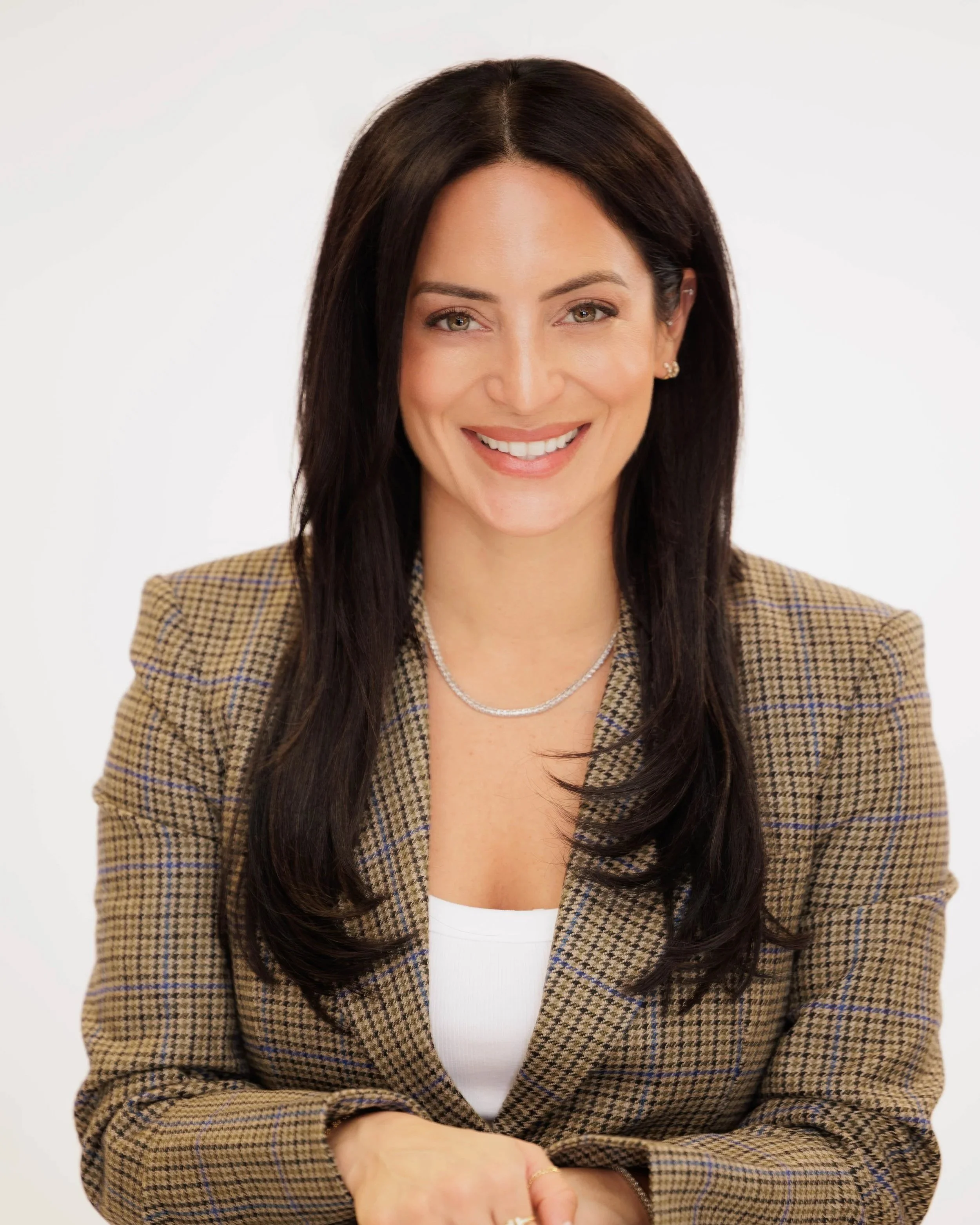 Portrait of a smiling woman with long dark hair, wearing a checkered blazer, white top, and jewelry, against a plain light background.