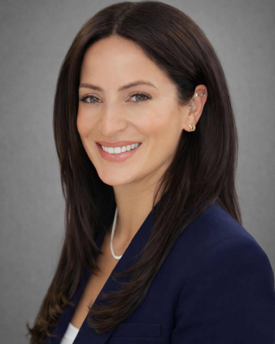 A professional woman with long dark brown hair, smiling, wearing a navy blazer, pearl necklace, and small earrings, against a plain gray background.
