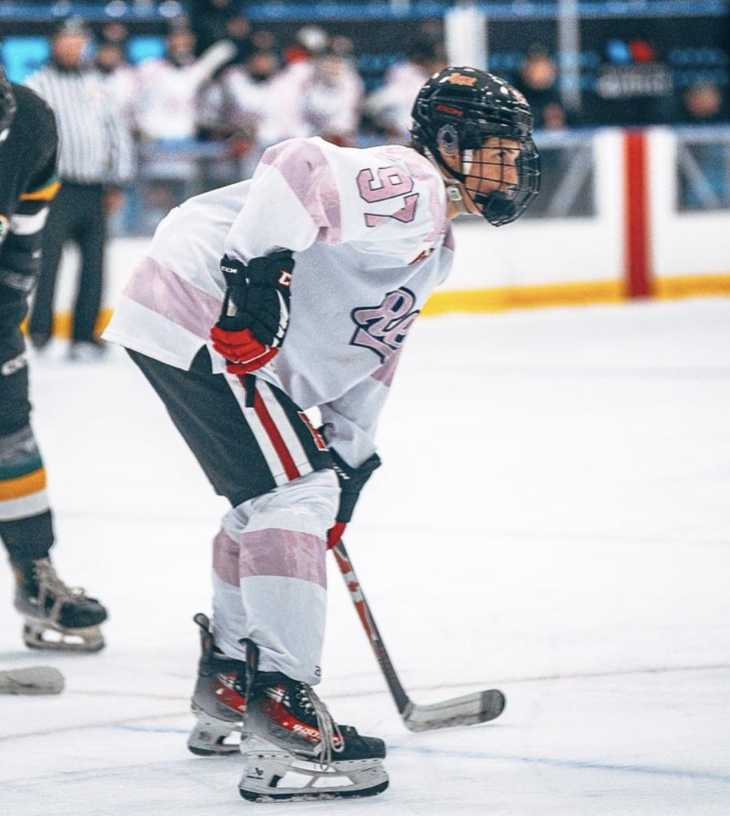 Hockey player in white jersey with pink accents, black helmet, and ice skates, leaning forward on the ice during a game.