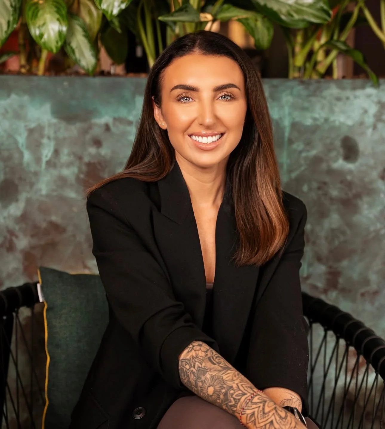 A woman with long brown hair, blue eyes, and a bright smile, wearing a black blazer, sitting in front of a green leafy background.