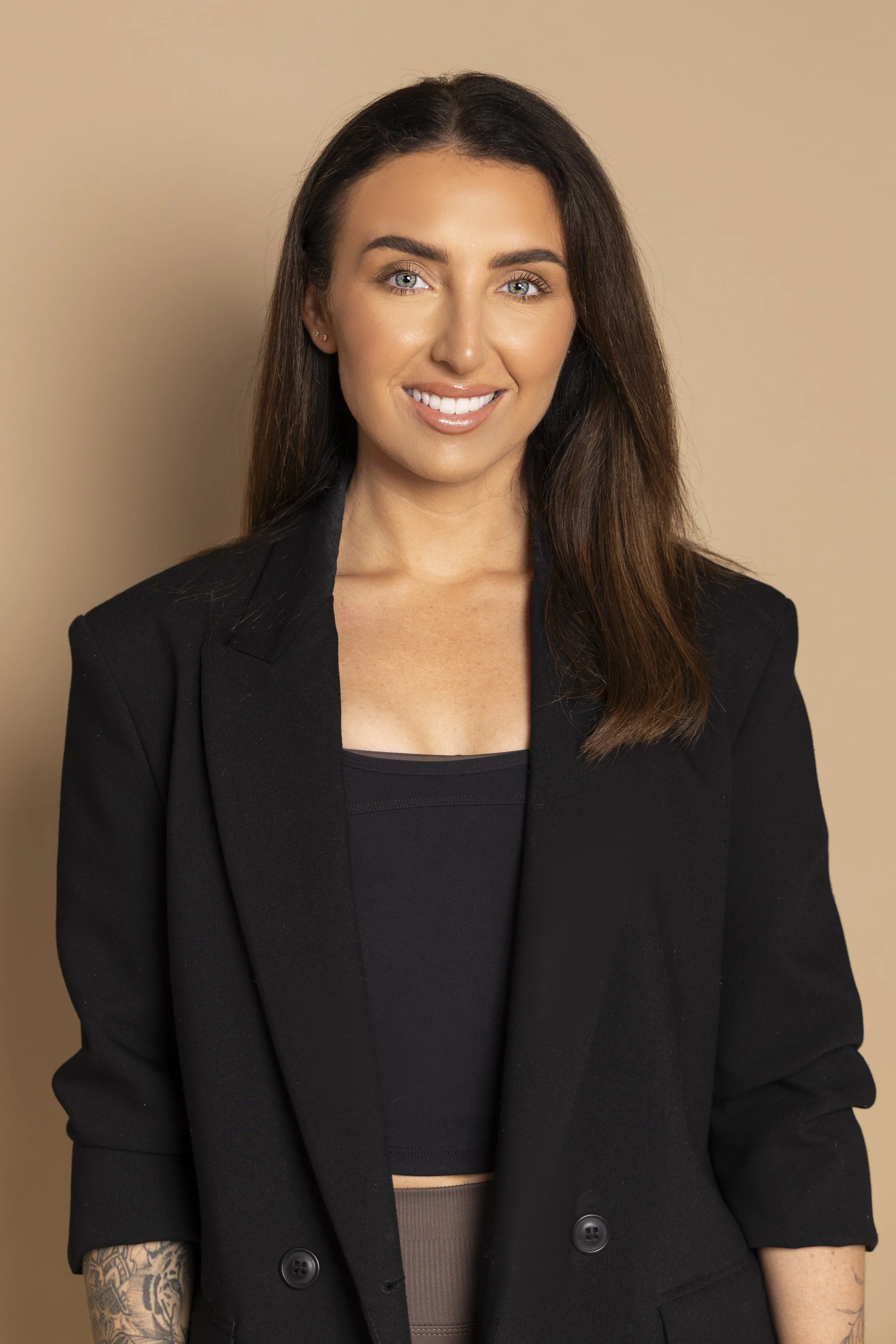 A woman with long brown hair, light skin, and blue eyes wearing a black blazer and black top, smiling in front of a beige background.