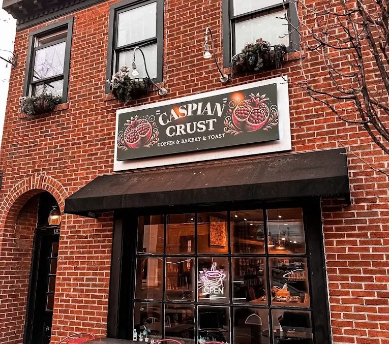 Exterior of a brick building with the sign 'Caspian Crust Coffee & Bakery & Toast' above the window, which has an 'Open' sign illuminated inside. The building has two small windows with flower boxes and decorative street lamps.