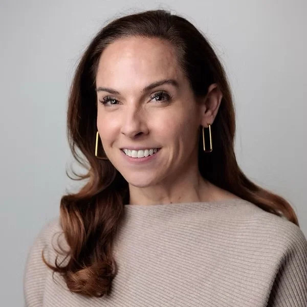 A woman with long brown hair, wearing a beige top and rectangular gold earrings, smiling and winking at the camera against a plain gray background.
