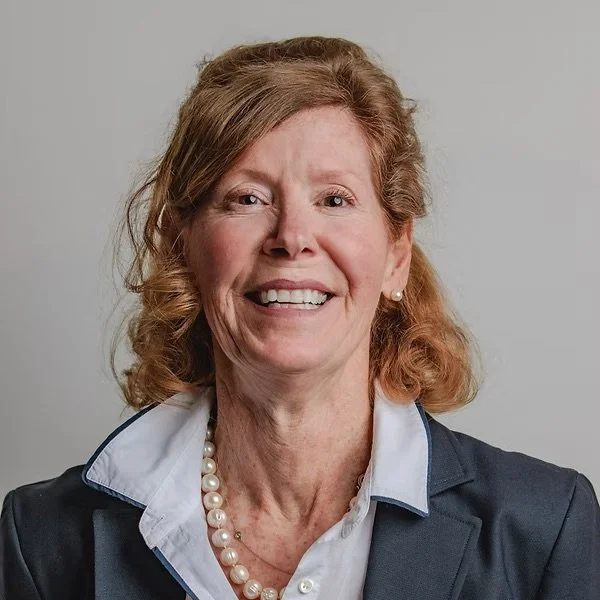 A smiling woman with curly red hair, wearing a pearl necklace, a white collared shirt, and a dark blazer, posed against a neutral background.