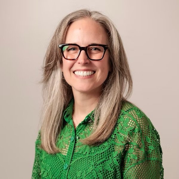 A woman with long gray hair, wearing glasses with black frames, and a green patterned lace blouse, smiling at the camera against a plain background.