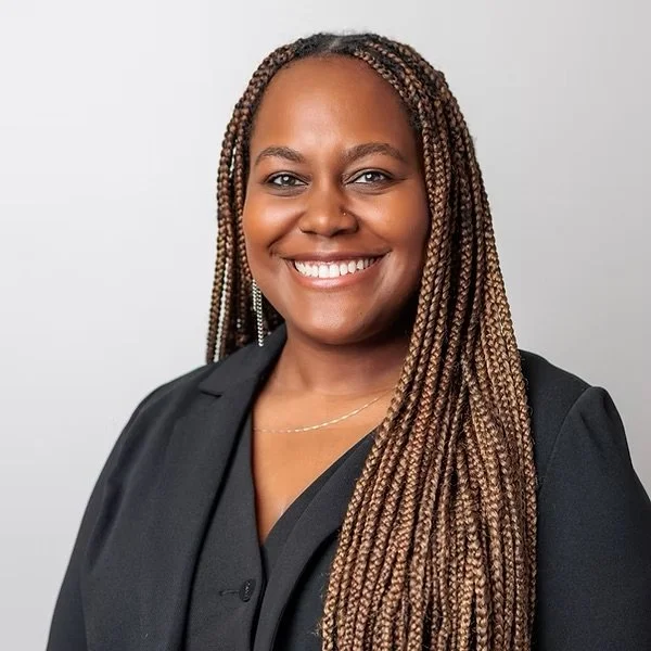 A woman with long braids smiling, wearing a black blouse, against a neutral background.