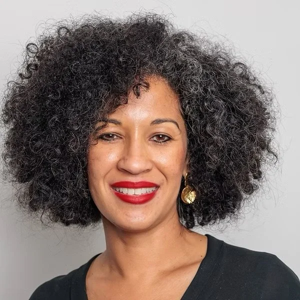 A woman with curly black hair smiling, wearing red lipstick, gold earrings, and a black top against a plain gray background.