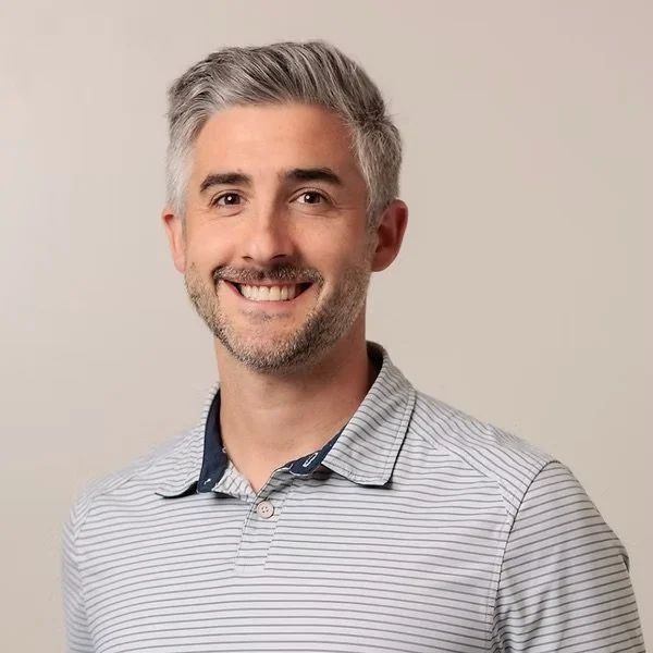 A smiling man with gray hair and a gray beard, wearing a light gray striped polo shirt, standing against a plain light beige background.