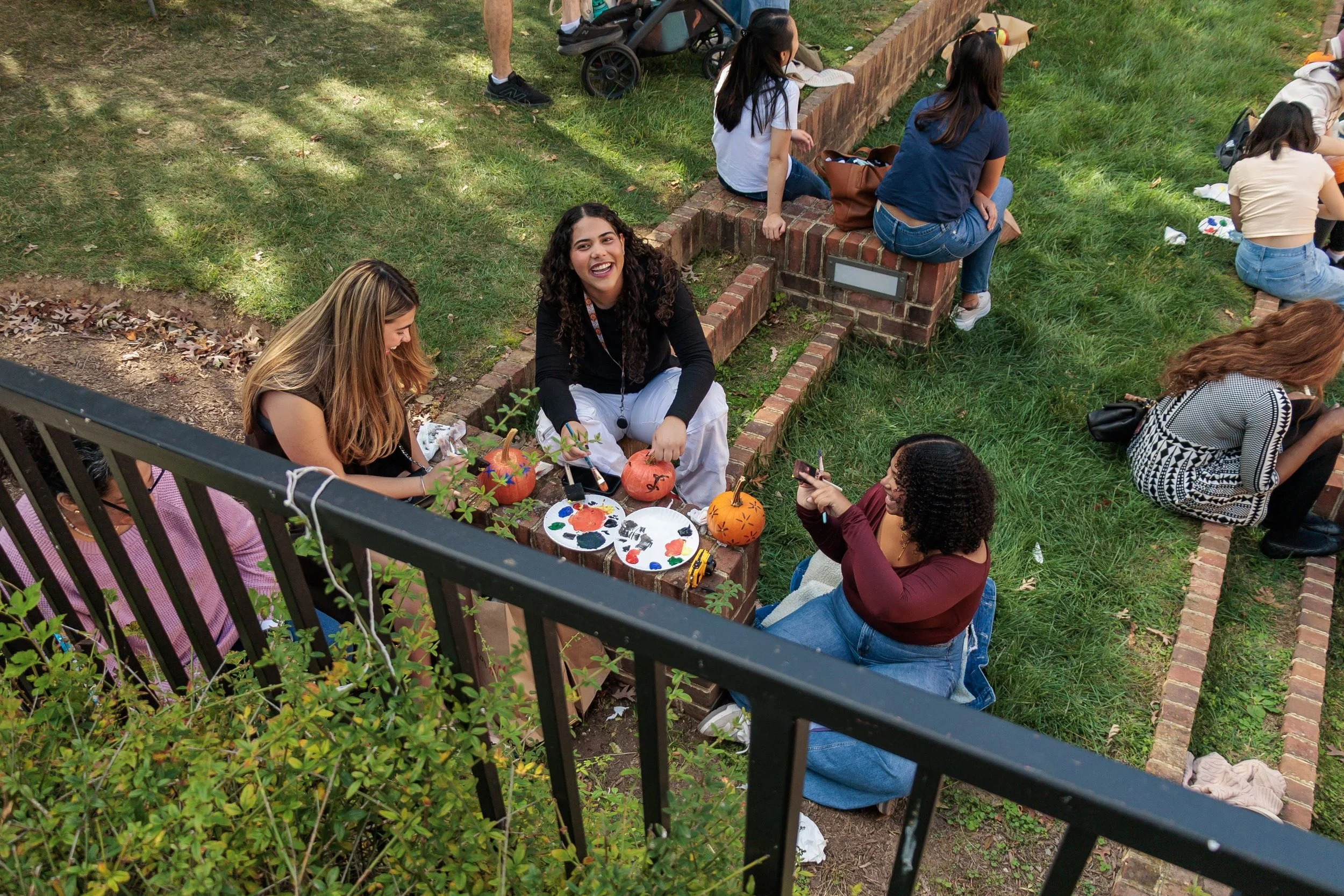 People sitting on a brick wall and ground in a park, painting Halloween pumpkins with festive decorations, with grass, trees, and a fence in the background.