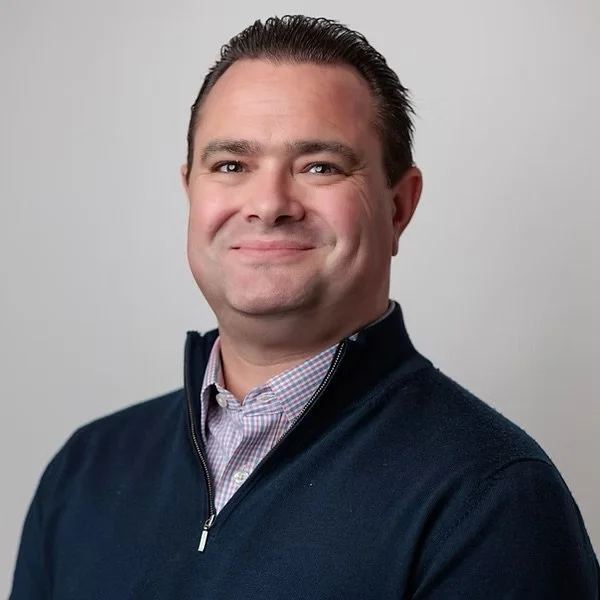 A professional headshot of a man with short dark hair, smiling, wearing a dark zip-up sweater over a collared checkered shirt, against a plain light gray background.
