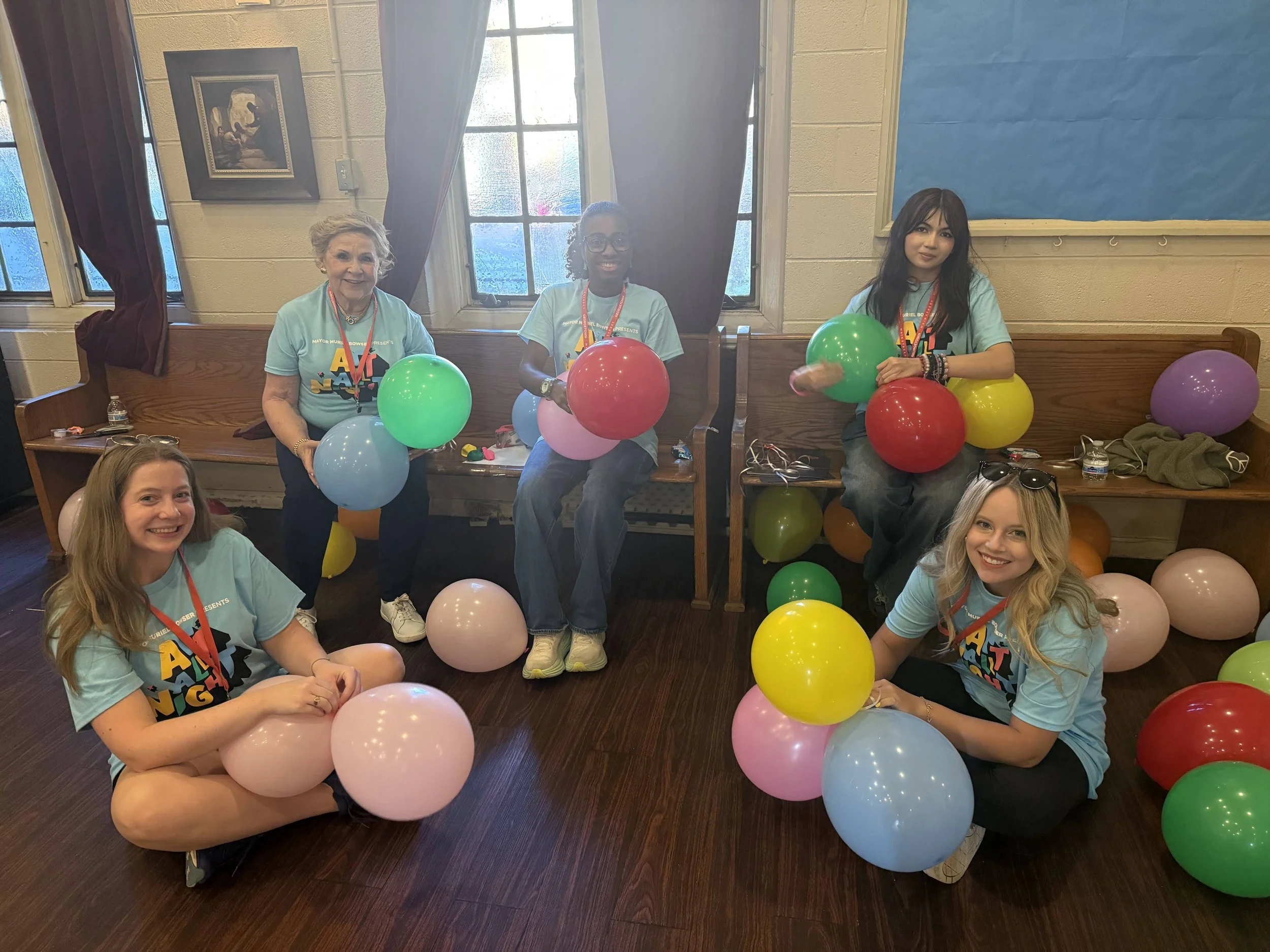 A group of five young women sitting on a wooden bench and on the floor, surrounded by colorful balloons, in a room with beige walls, windows, and a blue bulletin board, some smiling at the camera. All are wearing Art All Night t-shirts