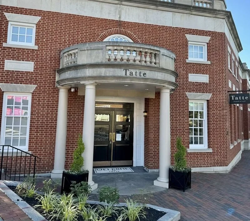 Brick building with white columns and a rounded balcony, storefront with the sign 'Tatte', potted plants, and a welcome mat at the entrance.