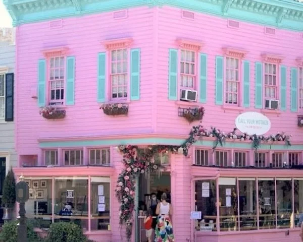 Pink building with teal shutters and window boxes, decorated with flowers and vines, featuring a large window display and a woman walking towards the entrance.