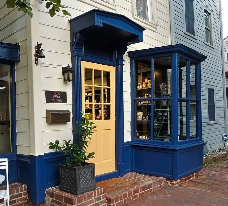 Exterior of Dent Place Market with a beige door, blue trim, a display window, and a potted plant outside.