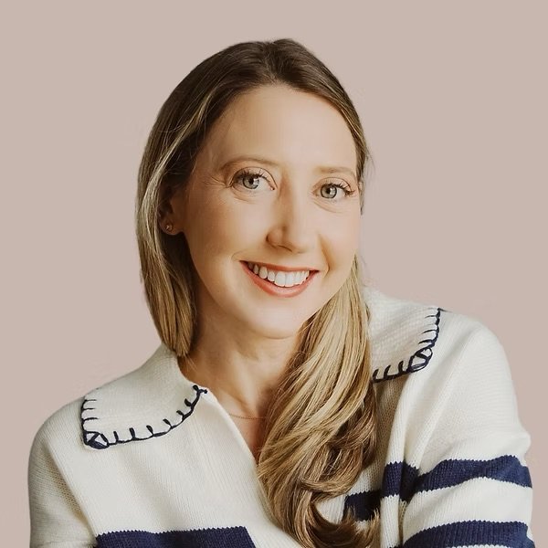 A woman with long brown hair smiling, wearing a white sweater with navy blue stripes and a scalloped collar, against a neutral beige background.