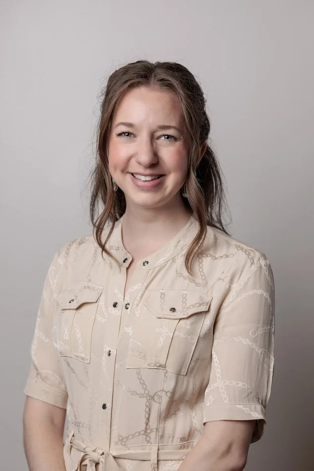 A woman with light brown hair, smiling, wearing a light-colored blouse with chain and pocket details, standing against a plain gray background.