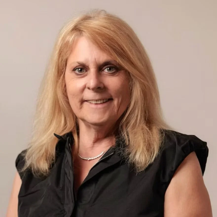 A woman with shoulder-length blonde hair wearing a black sleeveless top and a pearl necklace, smiling at the camera.