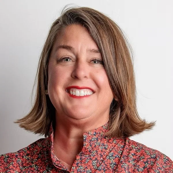 A woman with shoulder-length brown hair, smiling and wearing a red patterned top, against a plain white background.