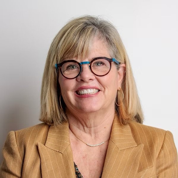 A woman with blonde hair, wearing glasses, gold hoop earrings, a pearl necklace, and a tan blazer, smiling against a plain white background.