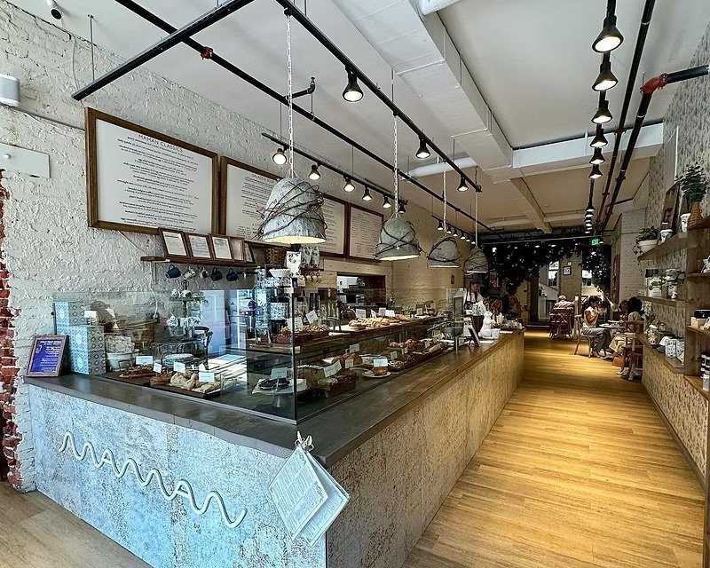 Interior of a modern bakery or cafe with a long display case filled with baked goods, pendant lighting, and customers seated at tables in the background.
