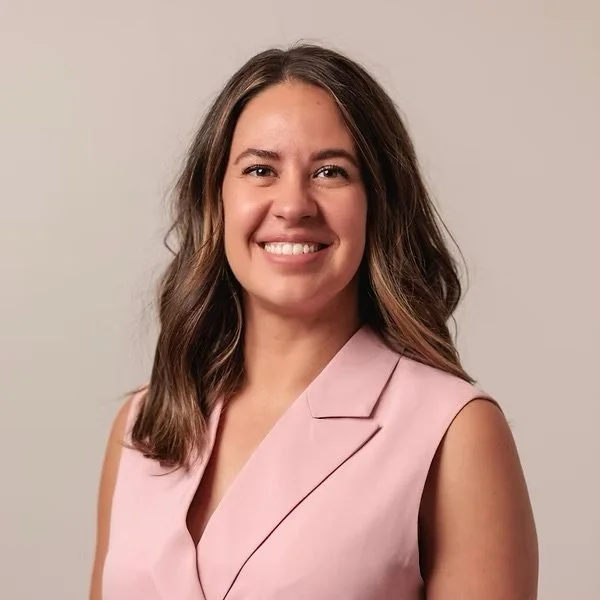 A woman with shoulder-length brown hair wearing a light pink sleeveless blazer, smiling against a plain light-colored background.