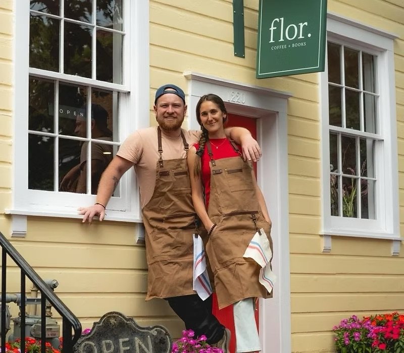 Two people, a man and a woman, standing outside a yellow building with white trim, wearing brown aprons. The man has his arm around the woman, and they are smiling. The building has a green sign that says 'flor' and 'coffee + books'. There are flowers at the base of the building and a black 'OPEN' sign near the steps.