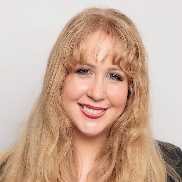 A smiling woman with long, wavy blonde hair and light makeup, wearing a dark top, standing against a plain white background.