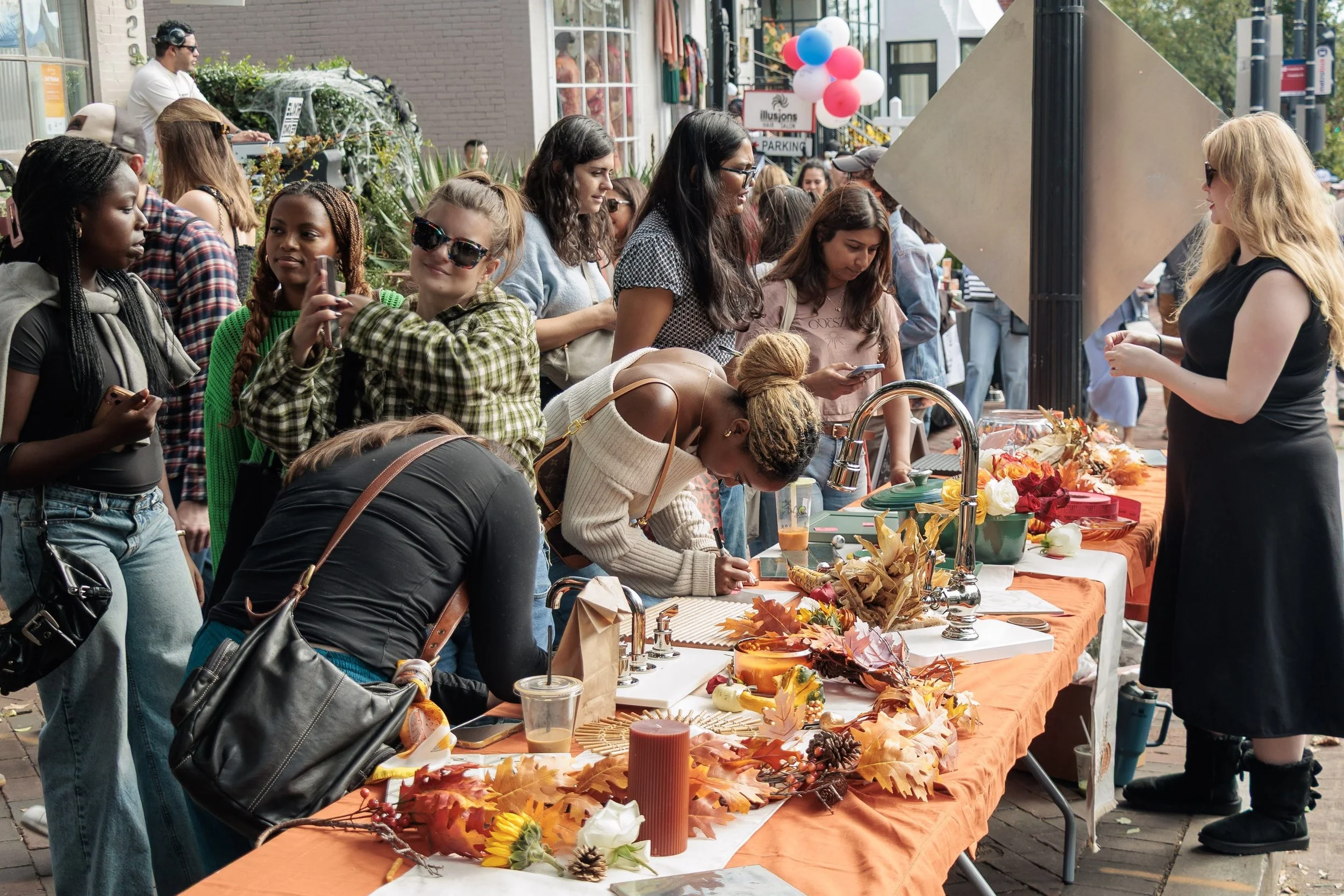 A photo of a diverse group of people on a sidewalk in Georgetown. There is a table will festive fall decor and two people appear to be writing. The atmosphere is lively and vibrant.