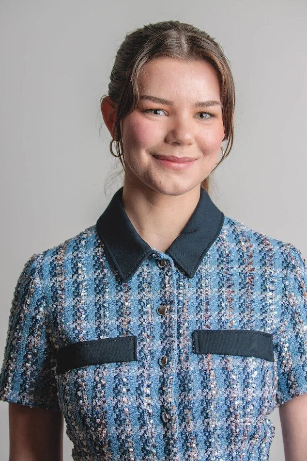 Young woman with brown hair tied back, wearing a blue patterned short-sleeve blouse with black collar and pocket accents, smiling at the camera against a plain gray background.