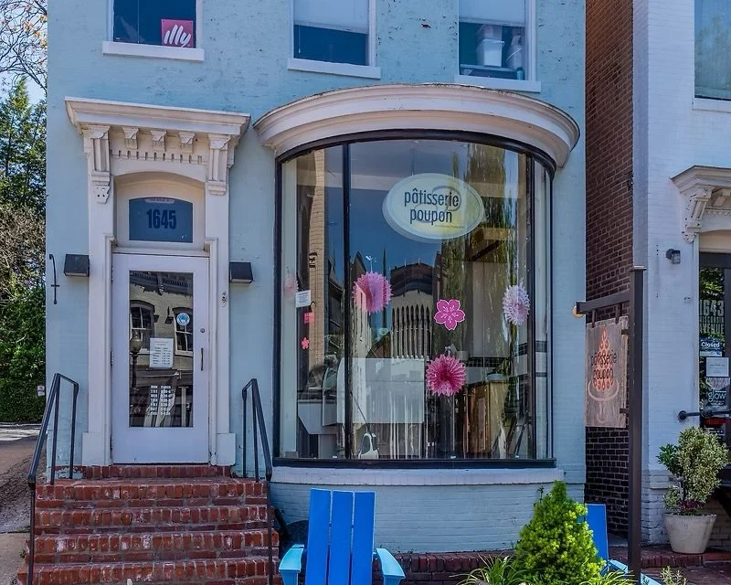 storefront with a curved glass window decorated with pink flowers and paper pom-poms, sign reading 'pâtisserie poupon', and white door with the number 1645