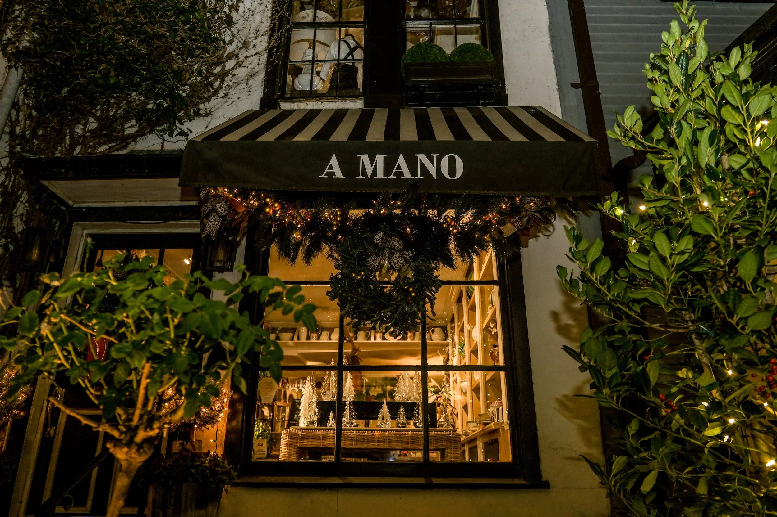 Storefront decorated with Christmas ornaments and greenery, with a black-and-white striped awning that has the words 'A MANO' written on it. Inside the store, Christmas trees and holiday decorations are visible.